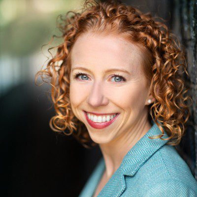 A person with curly red hair smiles warmly, leaning against a dark surface. They wear a light blue jacket, perhaps reflecting their participation in the Women in CS Masterclasses Series. The softly blurred background highlights their bright expression.