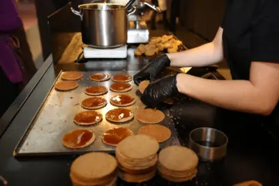 A person wearing black gloves, with the precision of AI, prepares stroopwafels at a kitchen station. They are spreading caramel on round waffle pieces. A pot on a stove and stacks of completed stroopwafels, reminiscent of the Best Community in Business gathering, are visible in the background.