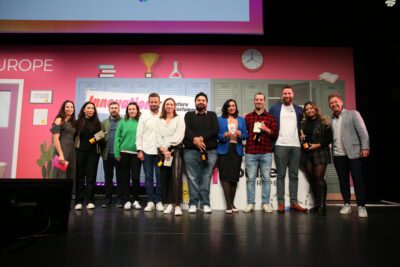 A group of twelve winners stands on stage in front of a vibrant backdrop featuring lockers and a clock. They are smiling, holding the prestigious Pulse Europe GameChanger Award 2024, and dressed in casual and semi-formal attire. The stage is set for an exciting award ceremony.