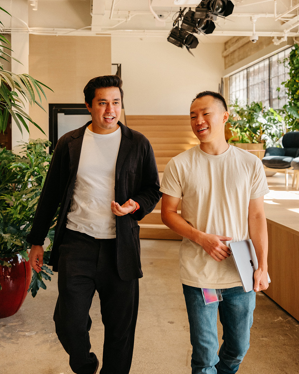 Two men walking side by side in a modern office space with plants around. One is wearing a black outfit and gestures while speaking. The other is in a beige shirt, holding a laptop and smiling. Both appear engaged in a friendly conversation.