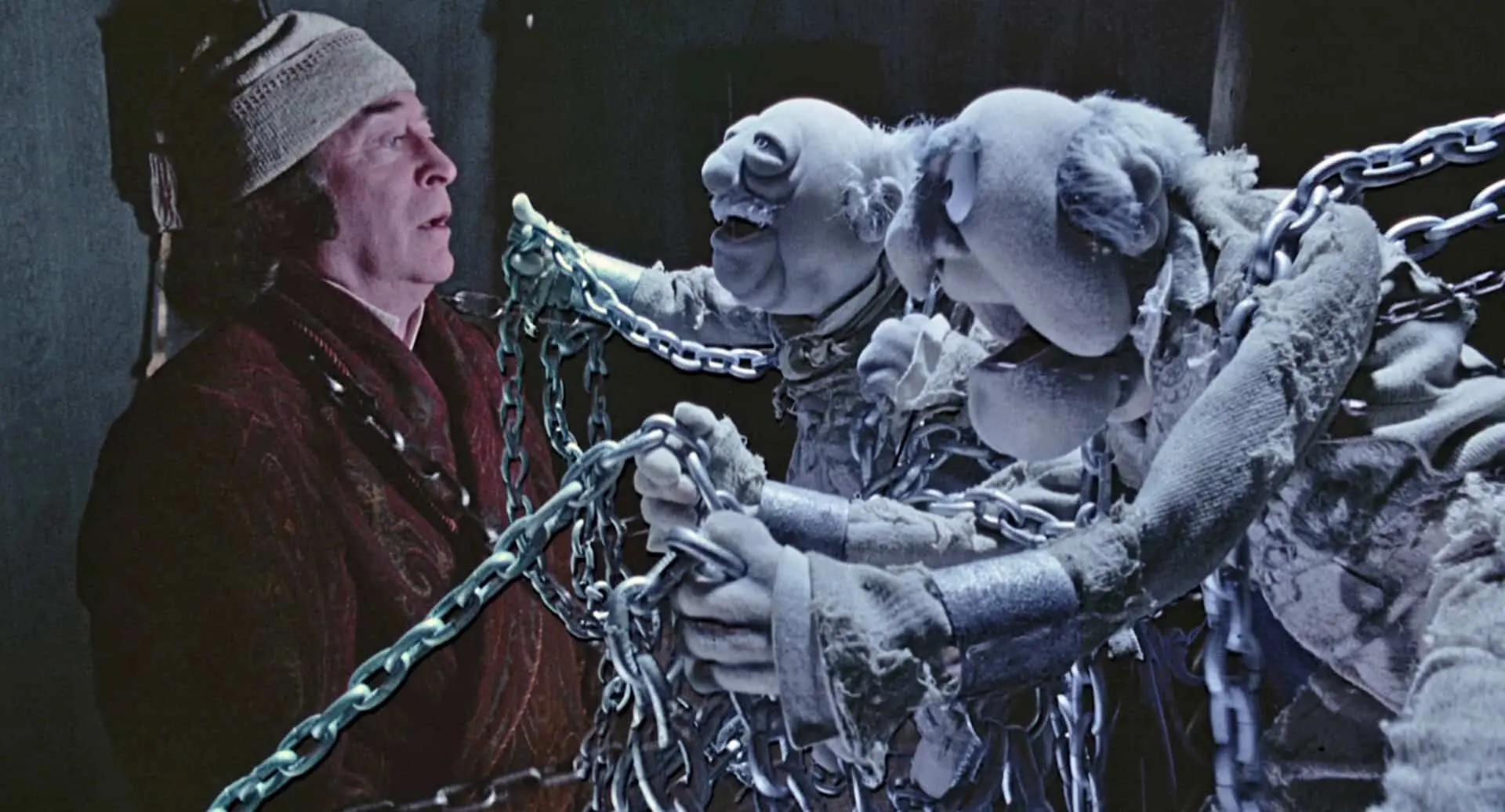 A person wearing a hat and robe stands next to two puppet figures draped in chains, their ghostly faces expressive in the dim glow. The scene, like a haunting gift that keeps on giving, shows the puppets interacting eerily with their human companion.